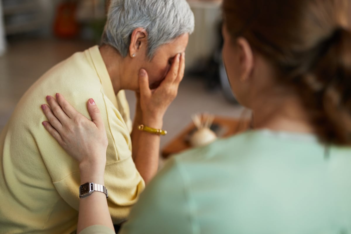 Idosa com o rosto coberto pelas mãos recebendo conforto, expressando a dor e o sofrimento emocional de quem sofreu um erro médico